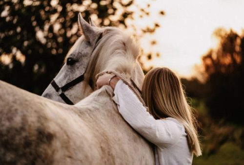 Blond young woman from behind embracing her white horse in nature in Majorca. Creative color editing with added grain. Part of a series.